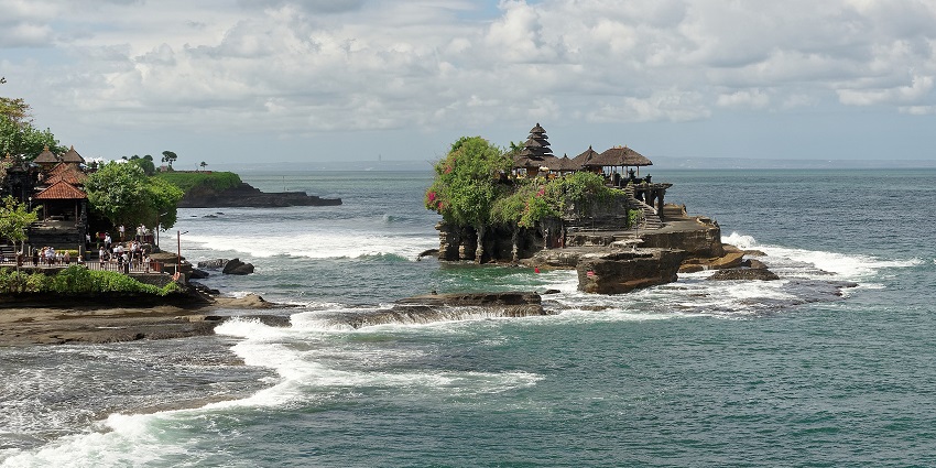 The Temple of Tanah Lot is perched on coastal rock, surrounded by ocean waves and rocky shore.