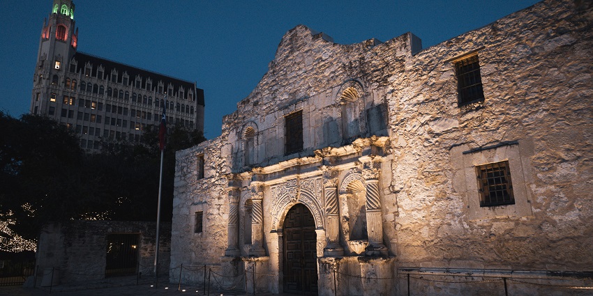 Historic Alamo mission in San Antonio lit up at night with a building in the background.