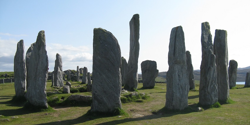Ancient standing stones, arranged in a circular pattern on grassy land under a bright sky.
