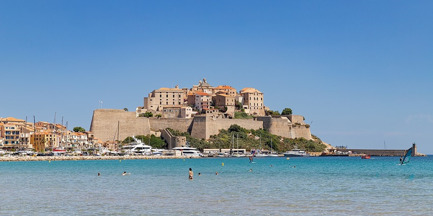 The citadel of Calvi with distant buildings, people in the sea and multiple boats parked.