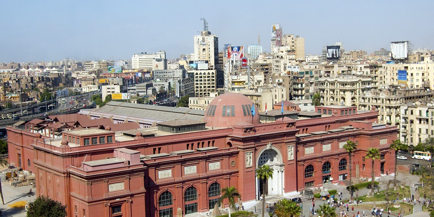 The Egyptian Museum in Cairo, with its red facade, is surrounded by city buildings and greenery.