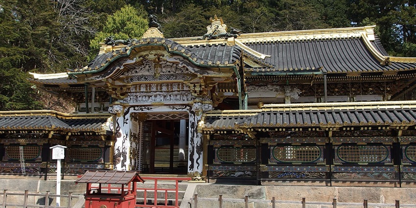 An ornate traditional gate with gold accents and intricate carvings, set against tall trees, is one of the most stunning historical places in Japan.