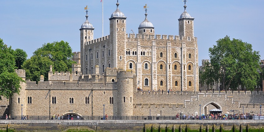 The Tower of London is seen from across the River Thames under a clear blue sky, with trees, one of the best historical places in London