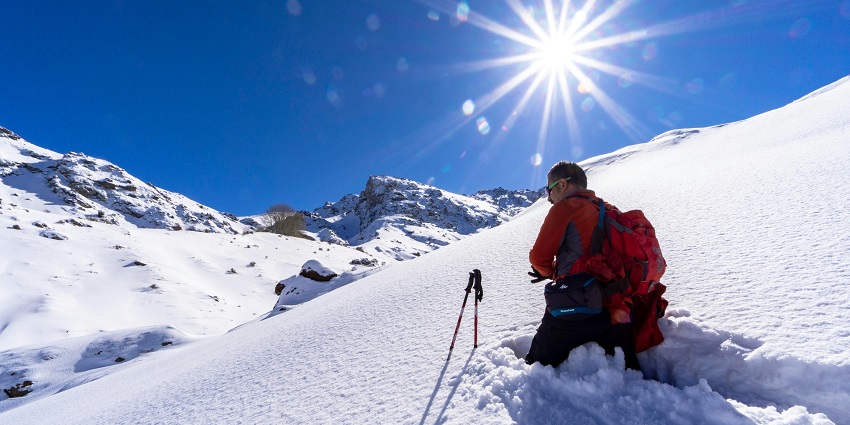 A backpacker hiking through snowy mountains under a clear blue sky, surrounded by snow-covered peaks and rugged winter terrain.