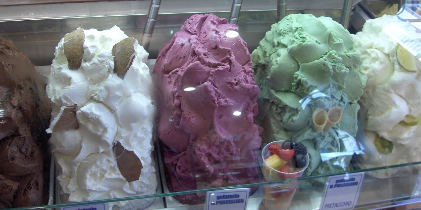 Cone with scoops of chocolate and pistachio gelato held against a blurred city street background.