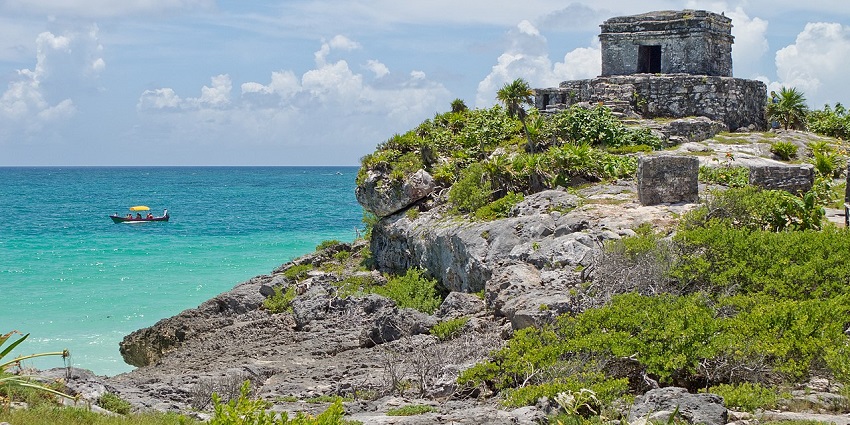 Tulum ruins with an old structure, a small entrance gate, green bushes and a boat on the sea