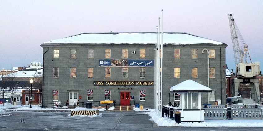 USS Constitution Museum in winter with snow, flags, and industrial cranes nearby.