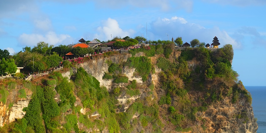 Uluwatu Temple is perched on a steep cliffside in Bali, overlooking the ocean on a sunny day.