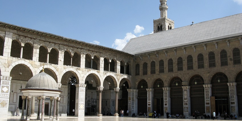 Umayyad mosque with a tall minaret and people sitting on the grounds, one of the historical places in Syria.