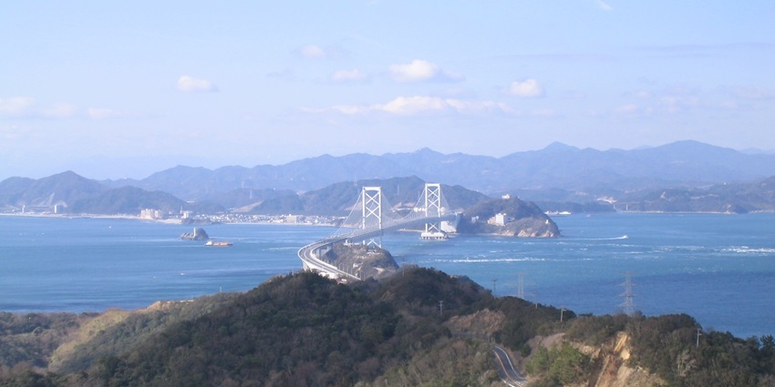 An image with coastal view of Shikoku Island with rocky shoreline, waves, and distant hills