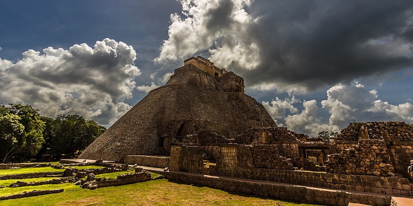 Uxmal with ruins, broken walls, a pyramid, green trees in the back and a cloudy sky