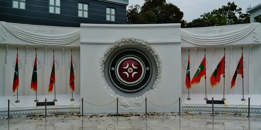 Victory monument in Malé with multiple Maldives flags, small fencing and carvings on the wall