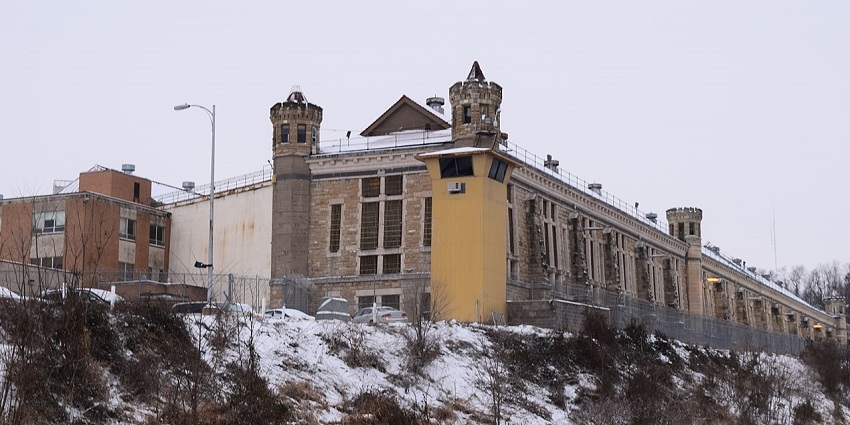 Historic Iowa State Penitentiary building with stone walls, barred windows, and watchtowers under a clear blue sky.