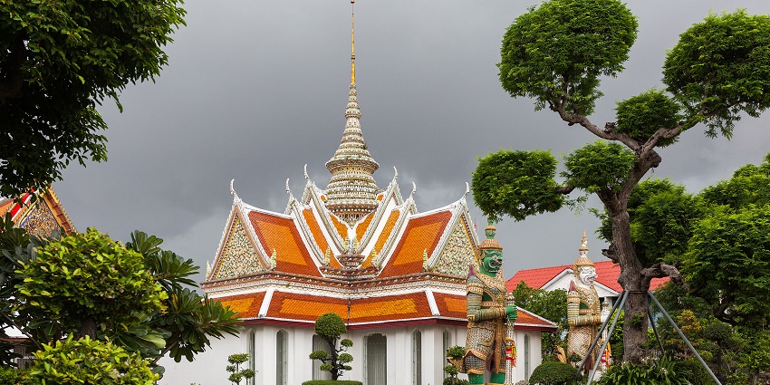An ornate Thai temple with an orange roof and guardian statues, surrounded by lush green trees, one of the most beautiful historical places in Thailand.