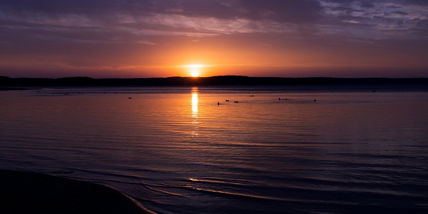 The sun sets over the ocean as people stand on the beach watching golden and orange sky colors.
