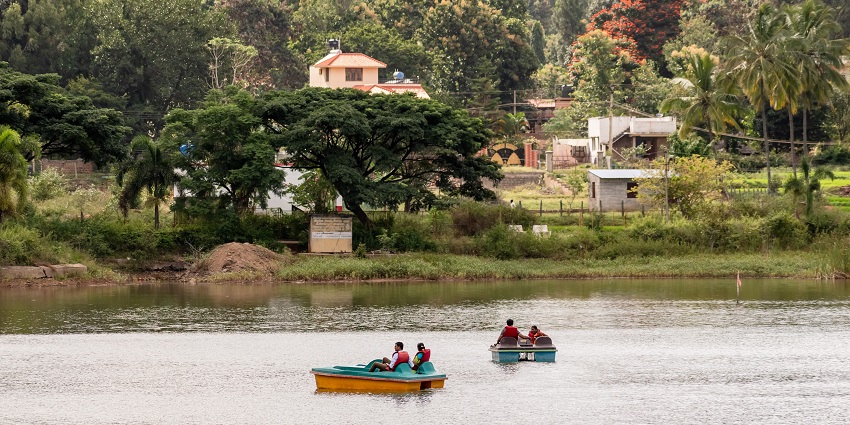 People boating on a calm lake surrounded by greenery and distant buildings under a sky.
