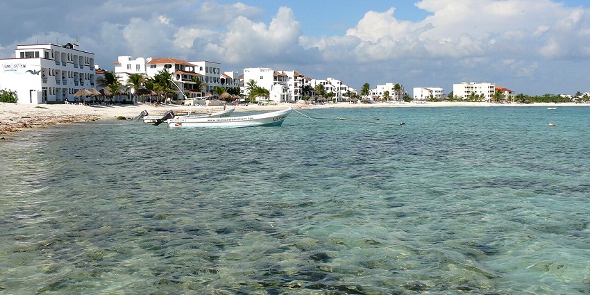 Sandy beach at Akumal’s Half-Moon Bay with gentle waves and small ships near the shore.