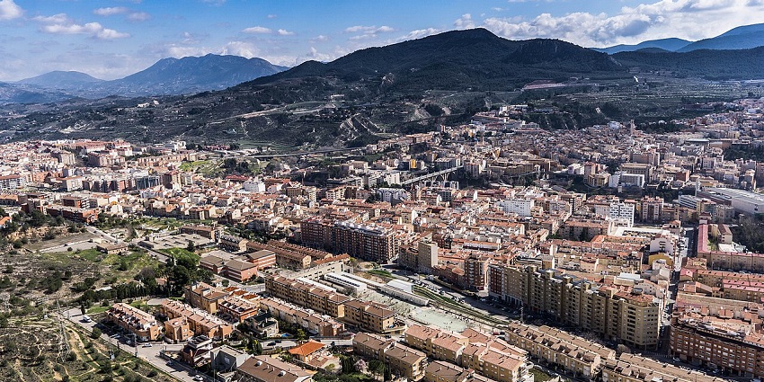 Alcoy city from above with multiple buildings, houses, and mountains under the blue, cloudy sky.
