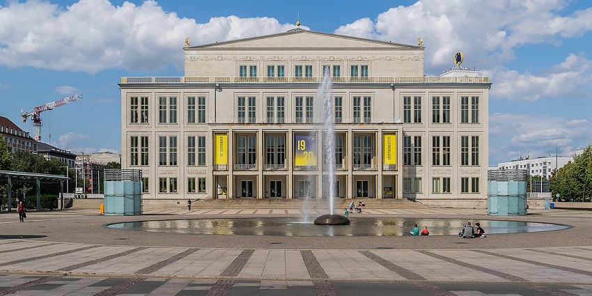 The Leipzig Opera House with tall windows and a stone facade, with a waterfall in front.
