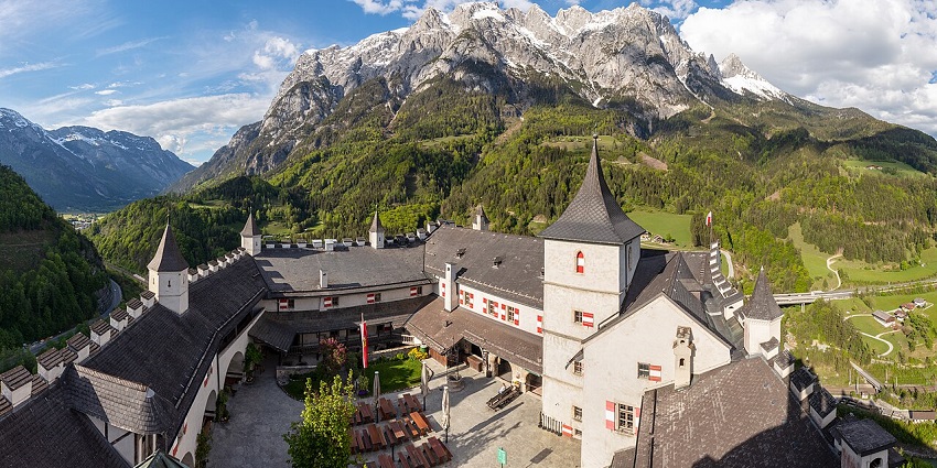 Hohenwerfen Castle in Austria sits atop a forested hill with mountains in the background.