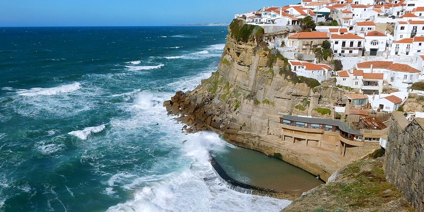 Waves crash against cliffs below white houses near Lisbon, one of the stunning places to visit near Lisbon.