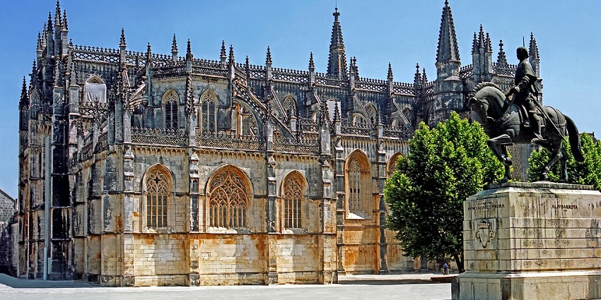 Gothic monastery with ornate spires and windows beside an equestrian statue in an open plaza.