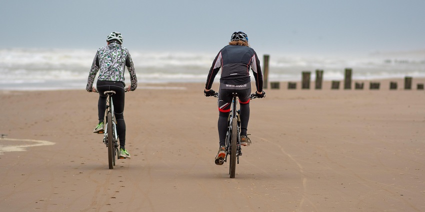 Two people cycling along a sandy beach with ocean waves and clear blue skies.