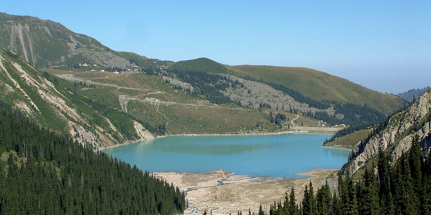 Big Almaty Lake with clear turquoise water surrounded by grassy slopes and greenery.