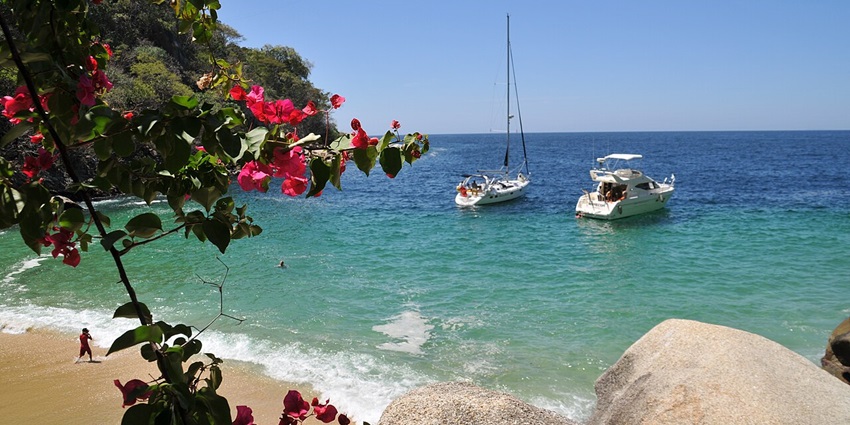 Two boats filled with people are cruising in the sea with the sandy coastline of the beach.