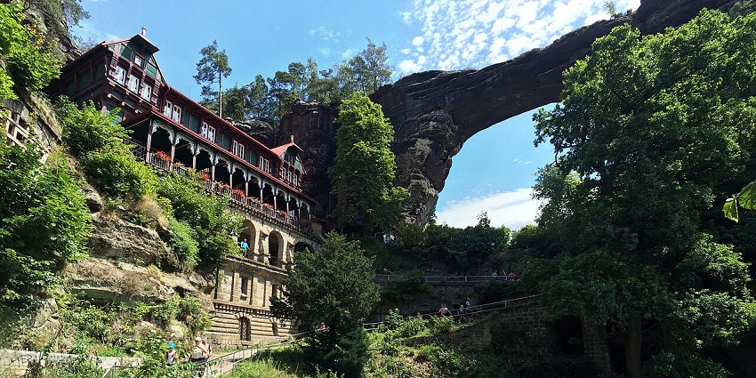 Scenic rock arch beside a rustic building nestled in lush greenery under a bright blue sky.
