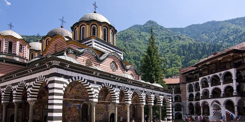 Rila Monastery in Bulgaria with striped arches, domes, and visitors in the courtyard.
