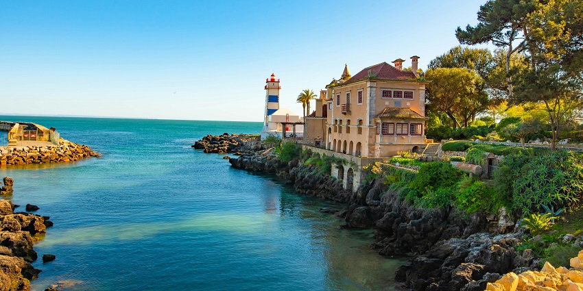A historic lighthouse stands near the sea with surrounding buildings under a clear sky in Cascais, Portugal.