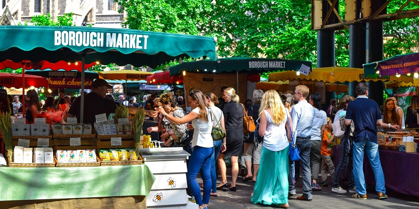 Food stalls at Borough Market in London with people browsing, under a clear sky.