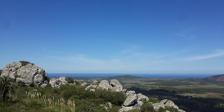 Cerro Pan de Azúcar with big rocks, dense green grass, ,distant grass plains and an ocean.
