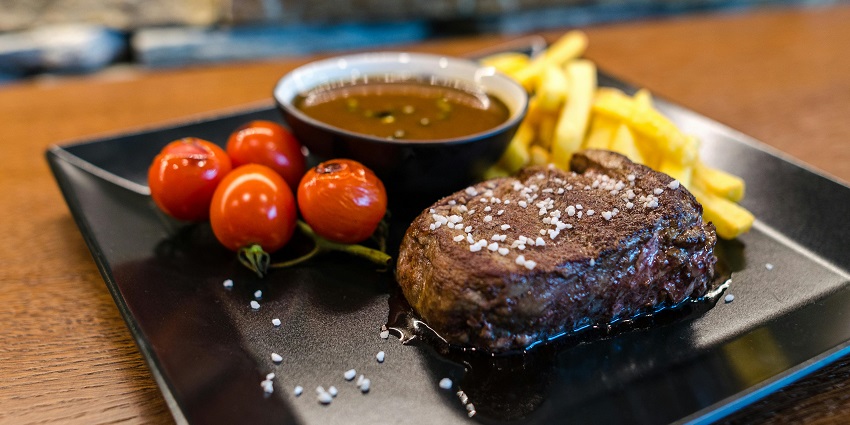 A plate of steak served with a brown gravy, French fries, and red fruits on a wooden table.