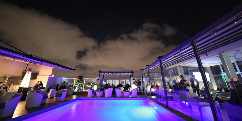 A pool in a rooftop lounge with purple lighting and people dining under a night sky.