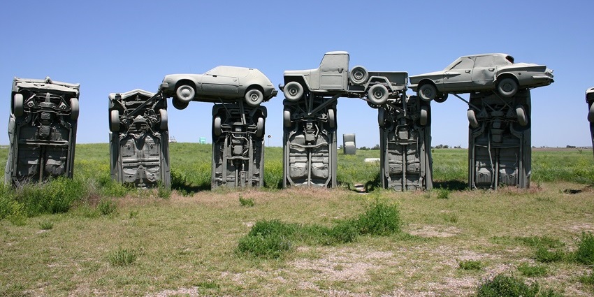A row of grey-painted cars stacked on each other under a bright blue sky on a dusty open ground.