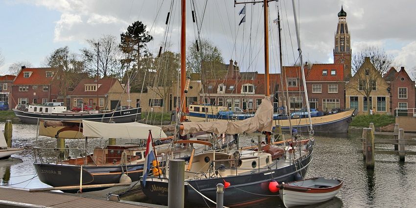 Sailboats docked in calm harbor waters with historic buildings under a blue, cloudy sky.