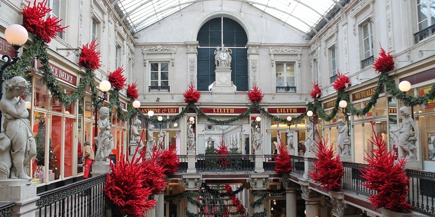 Elegant indoor gallery with festive red trees, garlands, statues, and a glass ceiling.