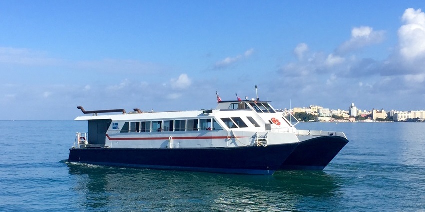 A white and blue passenger ferry cruising on calm blue waters under a clear, sunny sky.