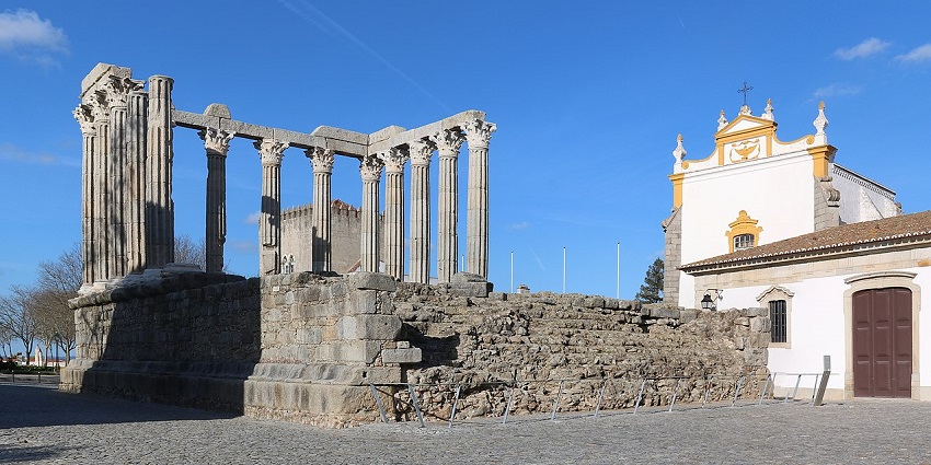 Ancient Roman temple ruins with tall stone columns beside a yellow church under a blue sky.