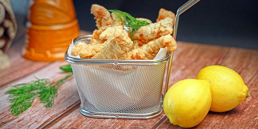 Fried fish in a small steel basket with two whole lemons on the side and small green leaves.