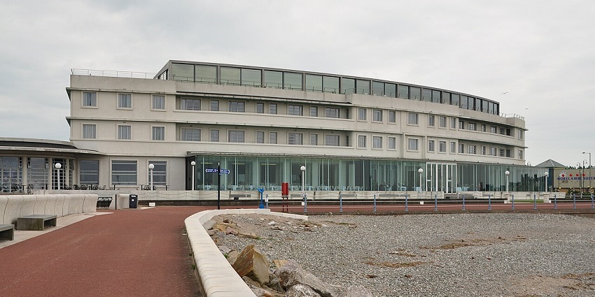 Modern curved beachfront building with large windows and a red walking path in front.