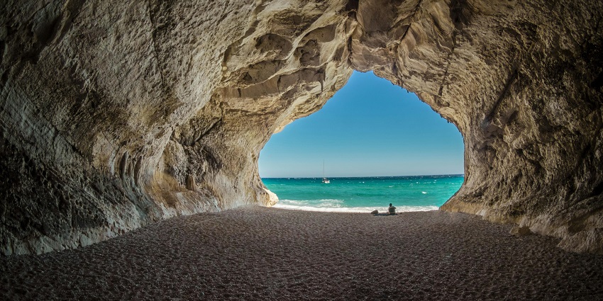 Inside a cave with unique rock formations, a person sitting at a distance on the beach.