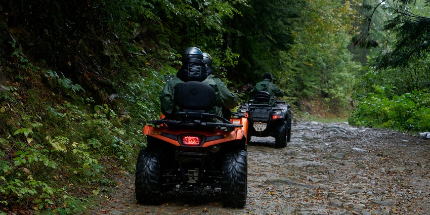 Two quad bikes with multiple people on the bikes and a dirt pathway surrounded by greenery.