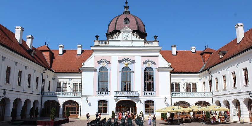 Elegant baroque-style palace with arched windows, red roofs, and people enjoying a sunny day.