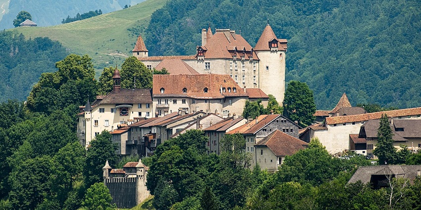 Hilltop castle complex with red-tiled roofs surrounded by greenery and forested mountains, one of the best places to visit near Geneva.