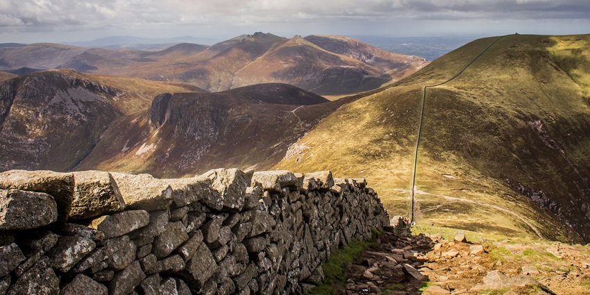 Hikers climb a misty mountain trail along a stone wall, surrounded by rugged green terrain.