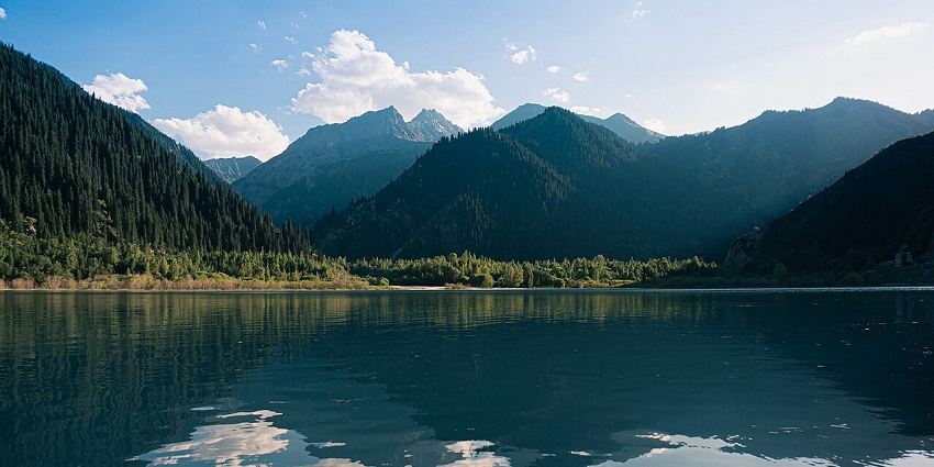 Issyk Lake with clear water reflecting clouds, surrounded by forested hills, one of the serene places to visit near Almaty.