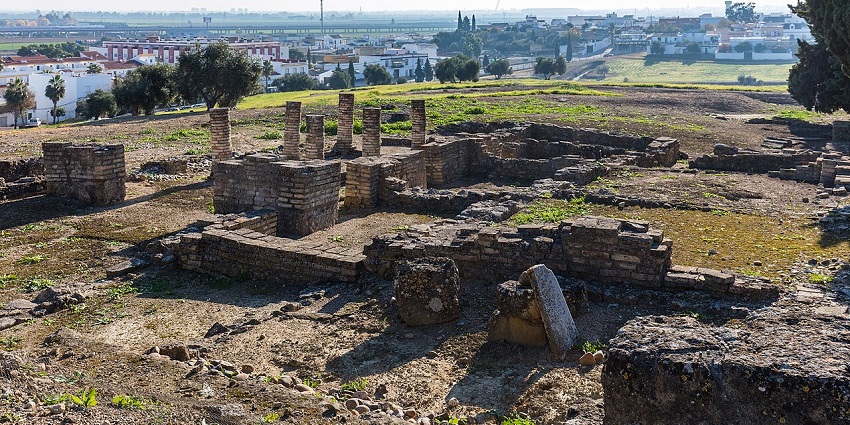 Ruin of ancient Roman city in Italica with broken pillars, walls, grass and buildings at a distance.
